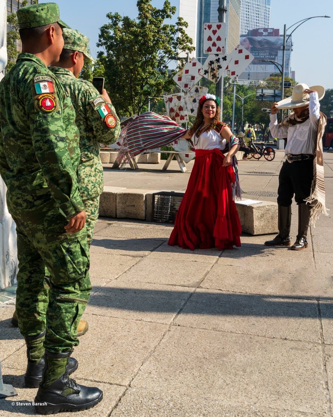 Two soldiers watch a woman in a red skirt and a man in a cowboy hat perform on a city sidewalk.
